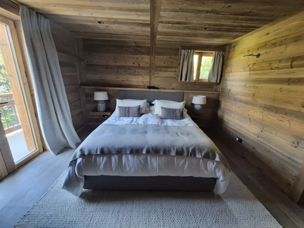 Rustic chalet bedroom with grey upholstered bed, ceramic table lamps and woven rug under a timber ceiling