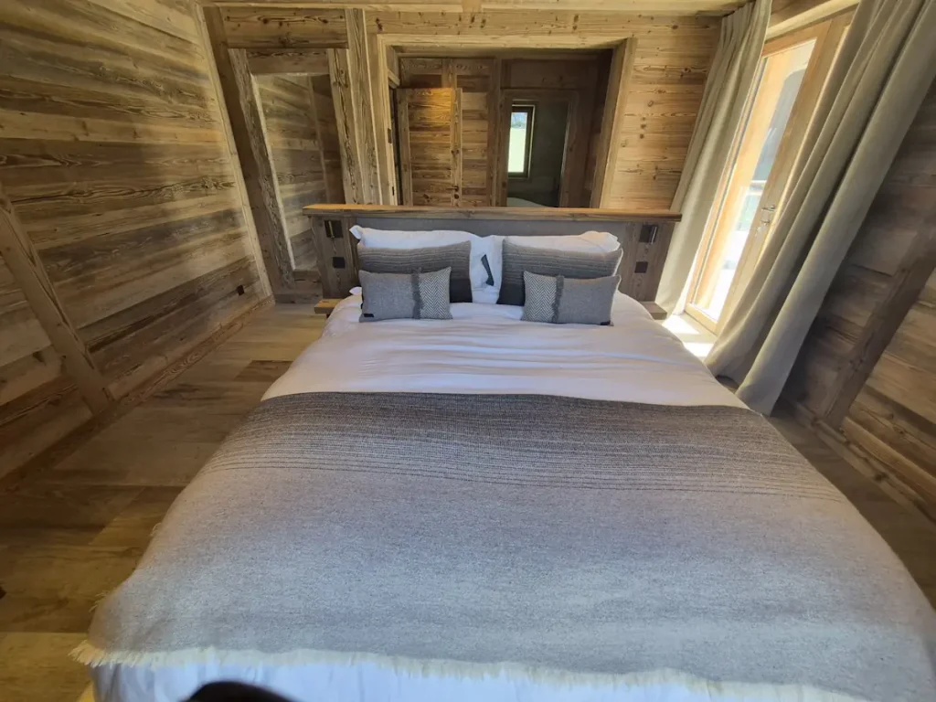 Timber-clad chalet bedroom with grey wool throw, herringbone cushions and balcony doors with linen curtains